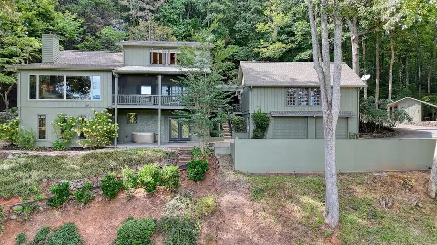 Two-story green board-and-batten main house nestled among mature hardwoods, viewed from the gravel driveway