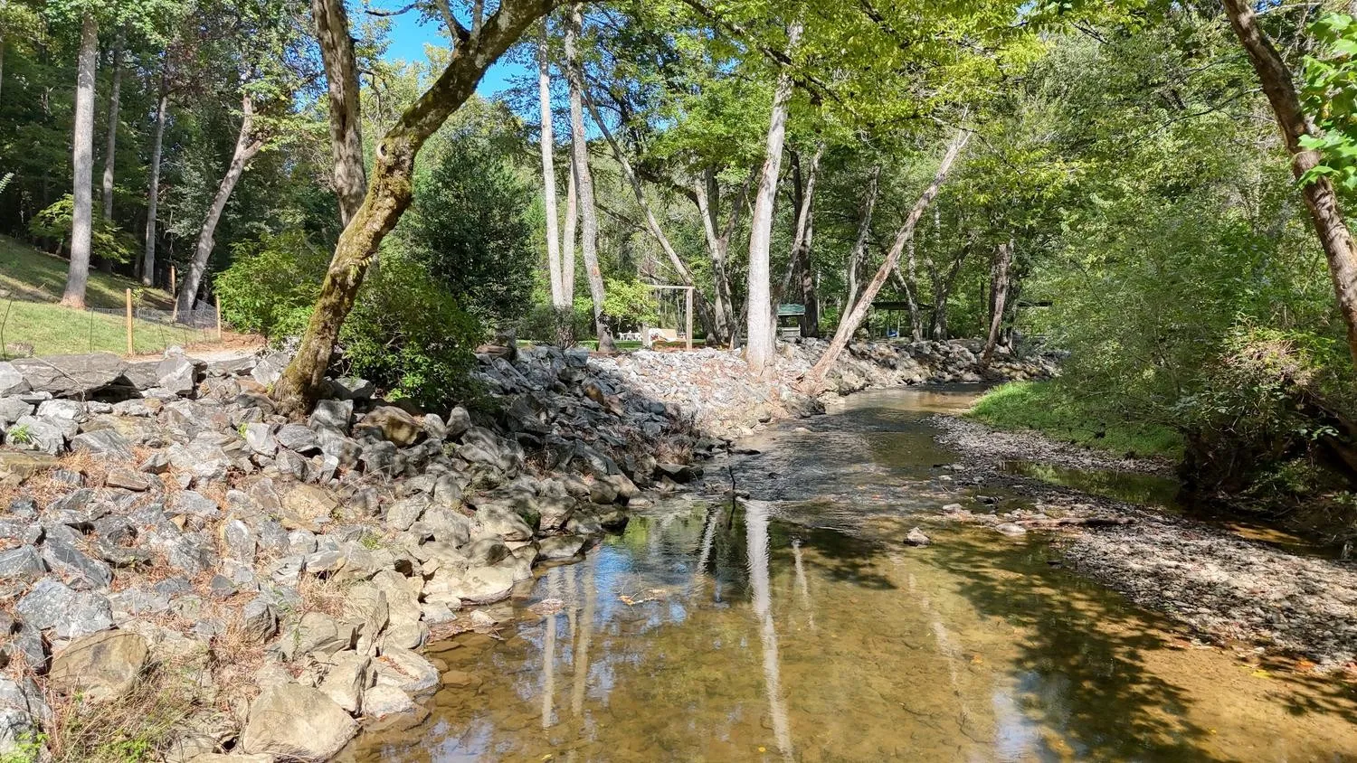 Brasstown Creek lined with rock banks and shaded by mature trees, with a pavilion and lawn visible in the distance