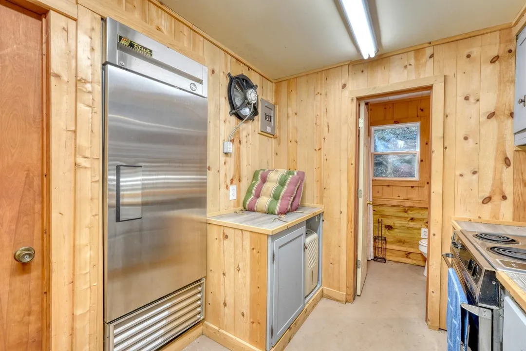 Knotty-pine prep kitchen with a full-height commercial refrigerator, electric range and counter space, opening to a restroom
